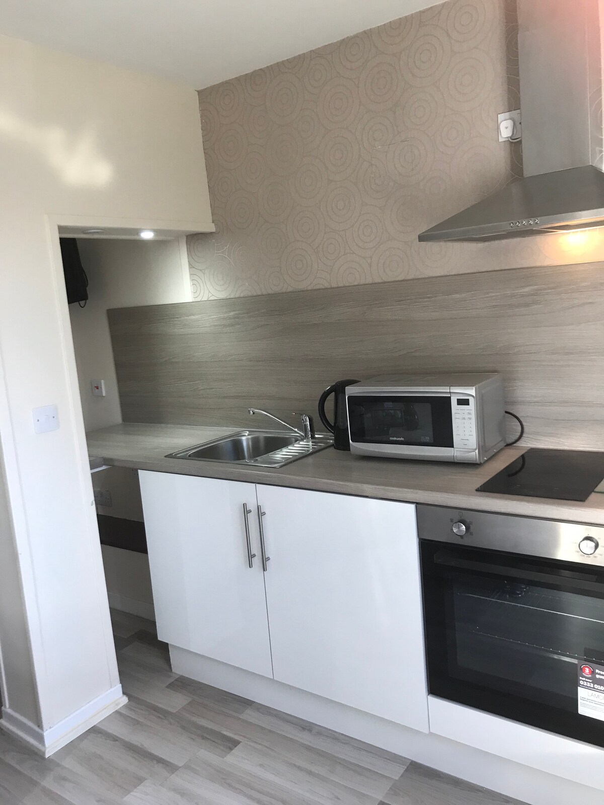 The kitchen features modern cabinetry with a light-colored finish. A stainless steel sink is situated under the wall-mounted cabinets, accompanied by a microwave and an oven. A neutral-toned backsplash complements the sleek countertop, while a bright light source enhances the space.