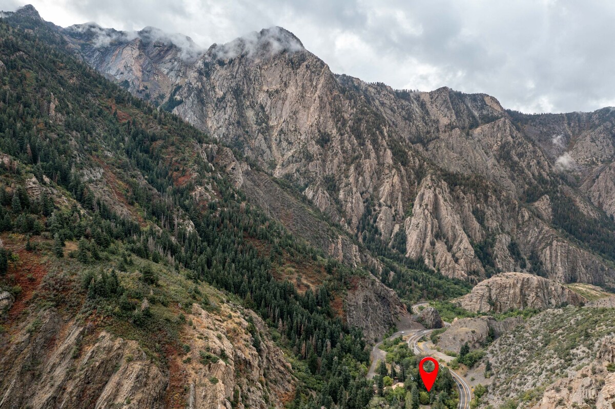 A sweeping view of rugged mountains rises majestically against a cloudy sky. Lush greenery adorns the slopes, contrasting with the rocky terrain. A winding road is visible in the lower part of the image, showcasing a remote and tranquil natural setting.