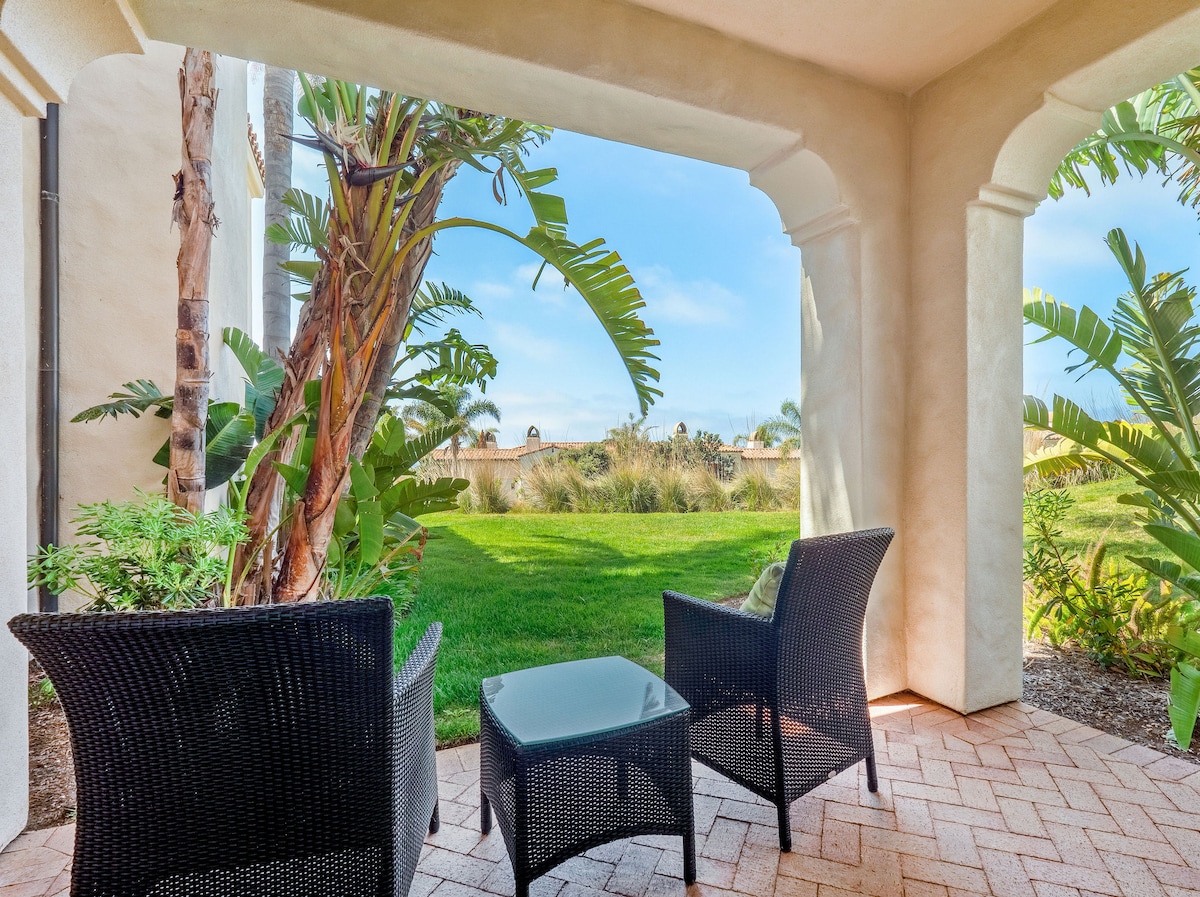 An outdoor seating area features two wicker chairs and a small table, surrounded by lush greenery. The space offers views of a grassy area, providing a peaceful setting for relaxation.