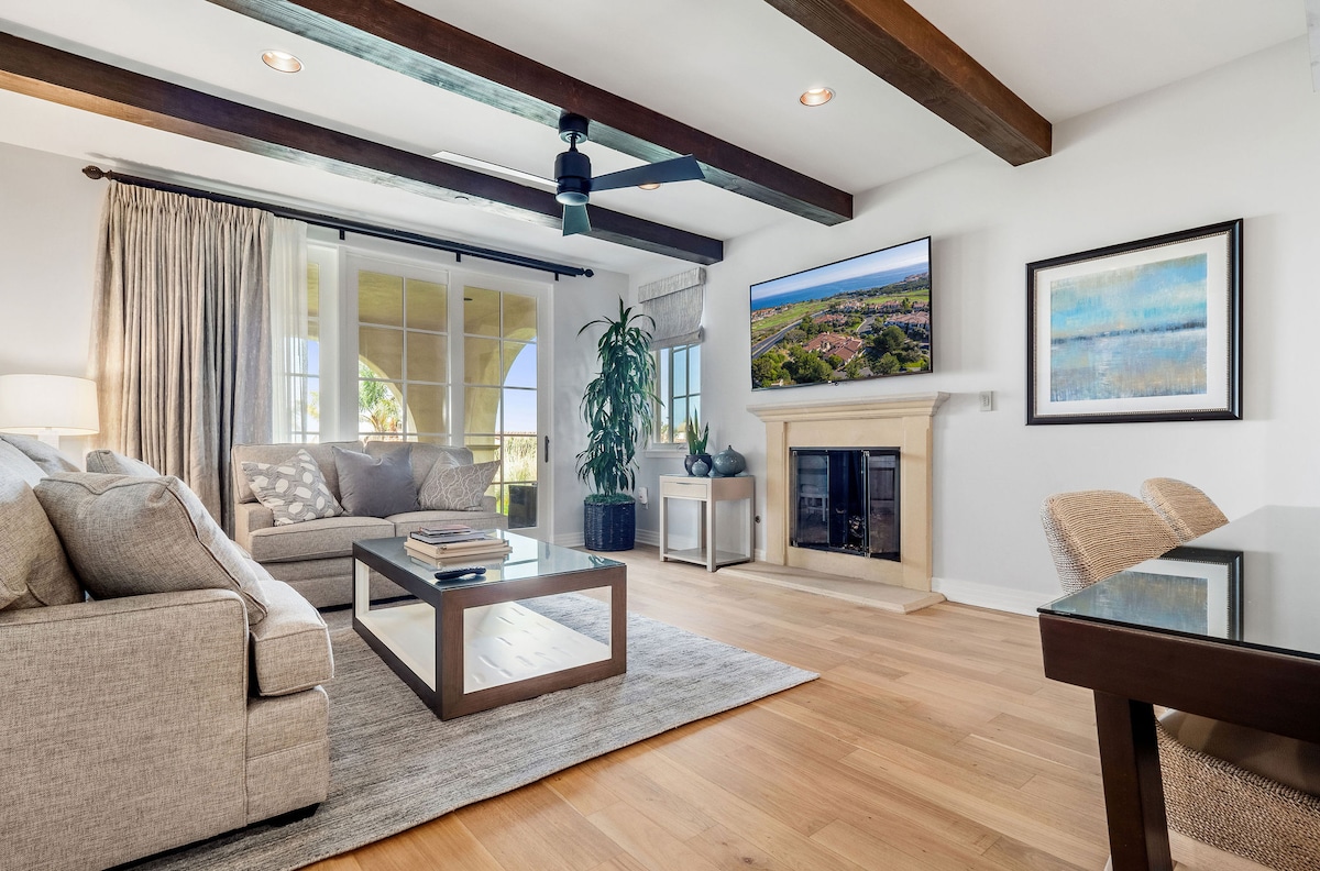 The spacious living room features natural wood beams overhead, a comfortable sofa, and a coffee table. A 60" flat-screen TV is mounted above a fireplace. Large glass doors provide access to the patio and allow plenty of natural light to fill the room.