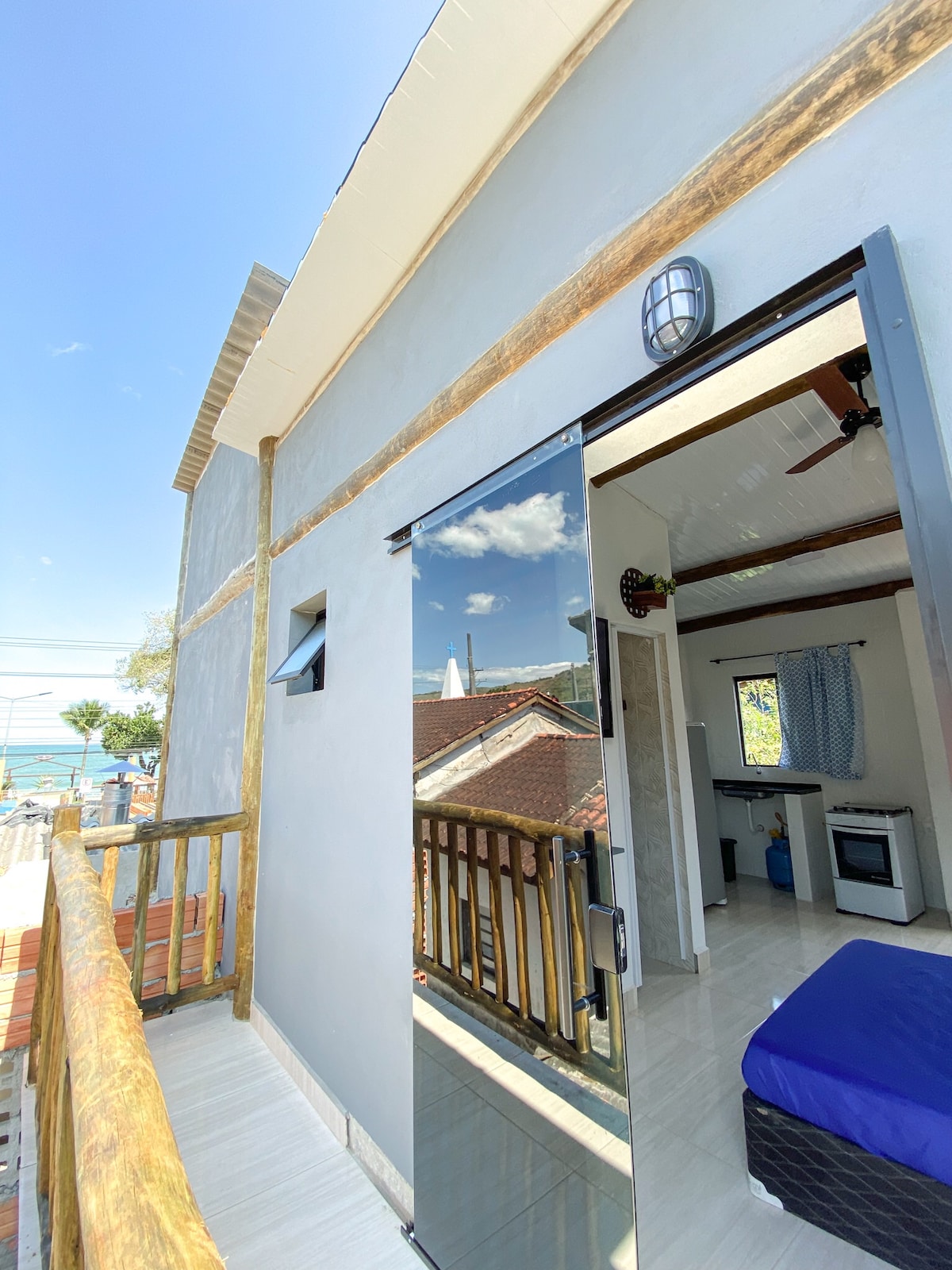 A modern exterior view features a glass door leading to a balcony with wooden railing. The backdrop displays a glimpse of the ocean, framed by a clear blue sky. The adjacent interior space is partially visible, suggesting a comfortable atmosphere.
