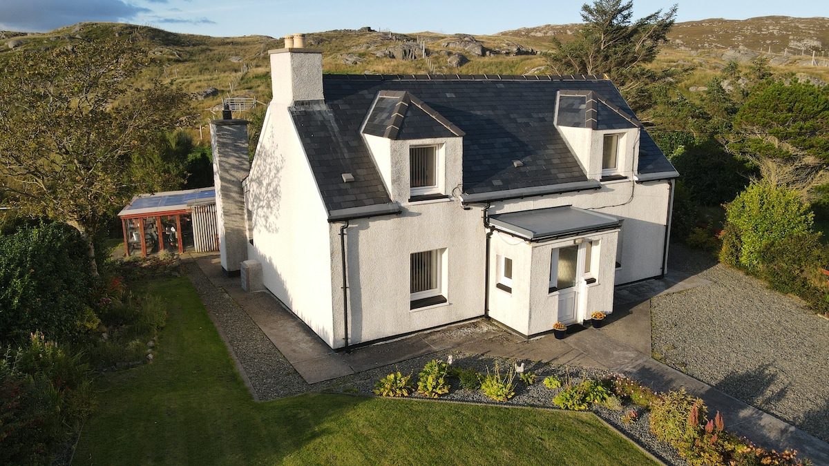 A charming two-story house features a white exterior with a slate roof. Surrounded by greenery, the property includes a well-kept garden and a gravel pathway leading to the entrance. A sunroom is visible on the side, enhancing the overall inviting atmosphere.