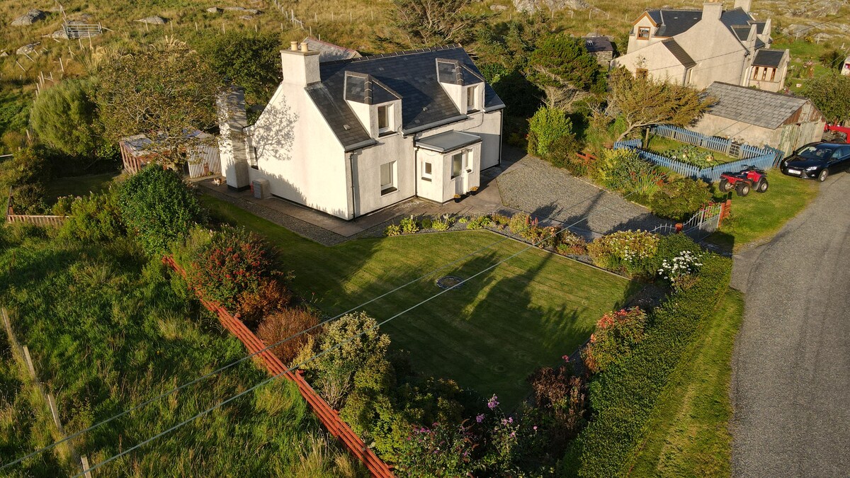 An aerial view of Taigh Chaluman showcases the charming croft house surrounded by a well-maintained garden featuring various shrubs and flowers. The property is bordered by a low wooden fence and offers a neatly trimmed lawn, with additional homes and greenery visible in the background.