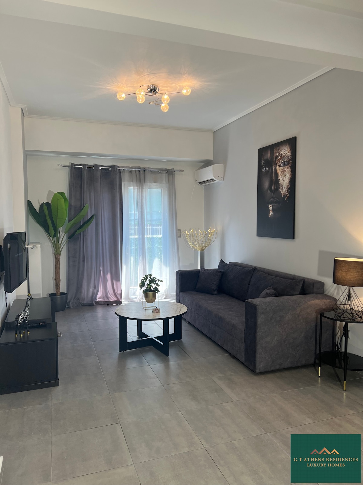 A modern living area is presented, featuring a dark sofa paired with a round glass-topped coffee table. Natural light filters through sheer curtains, complementing the neutral color palette. A decorative plant adds a touch of greenery, while a wall-mounted TV is positioned for convenient viewing.