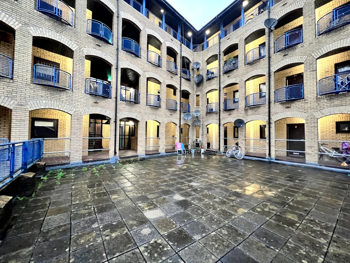 A spacious courtyard is surrounded by brick buildings featuring arch-shaped windows and balconies. The area is paved with large tiles and illuminated by soft lighting. A bicycle is parked along one wall, and colorful chairs are placed within the open space, creating an inviting communal area.