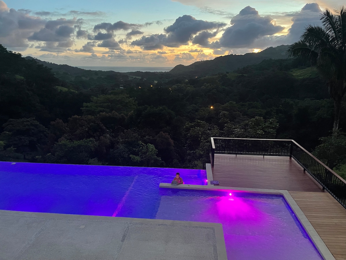An infinity pool extends toward the horizon, with gentle purple lighting accentuating the tranquil waters. Lush greenery envelops the area, while stunning views of the mountains and sky are captured during sunset, creating a serene outdoor space for relaxation.