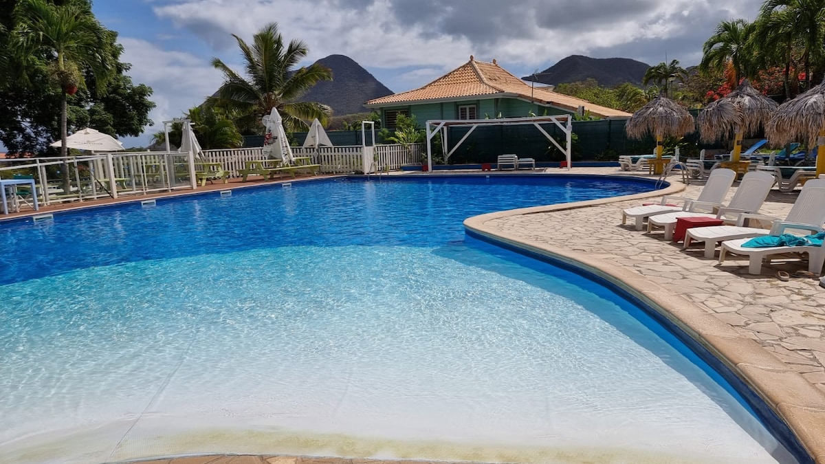 The outdoor pool is surrounded by lush palm trees and mountain views. Sun loungers and umbrellas are arranged around the pool deck, inviting relaxation. The shallow end of the pool features a gradual slope, enhancing accessibility for all guests.