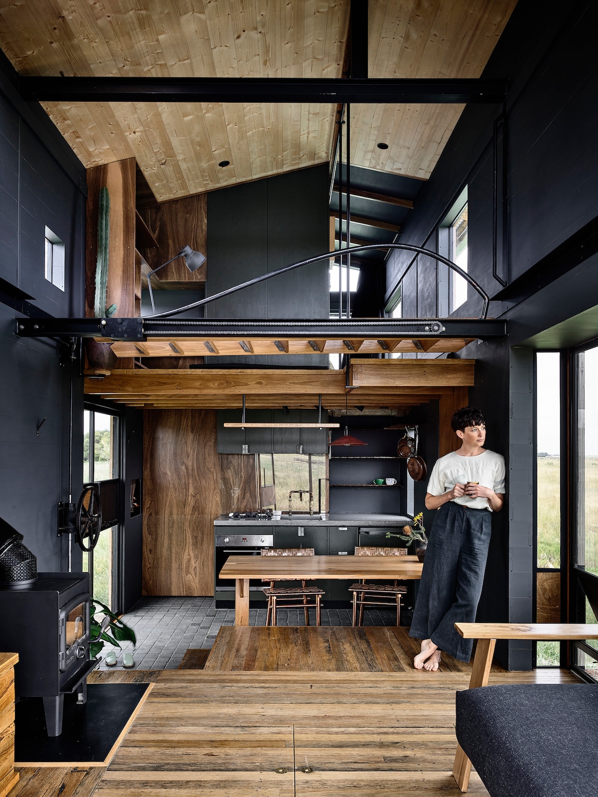 An open living space features high wood-paneled ceilings and large windows, allowing natural light to illuminate the interior. A kitchen area with modern appliances and a dining table is visible. A cozy woodfire stove stands in the corner, enhancing the warm atmosphere.