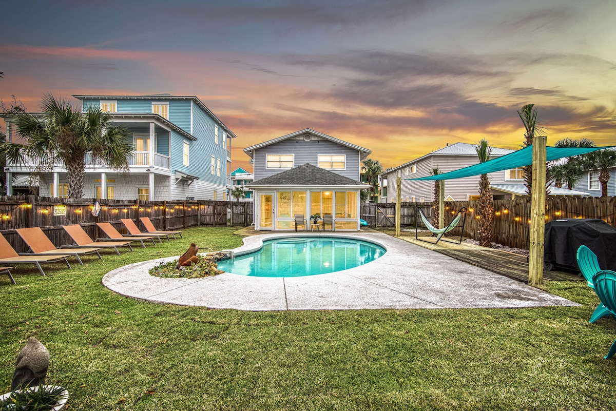 A large fenced backyard features a private pool surrounded by a spacious deck. Chaise lounges are arranged for relaxation under the evening sky. A hammock is positioned nearby, with lush green grass completing the serene outdoor atmosphere.