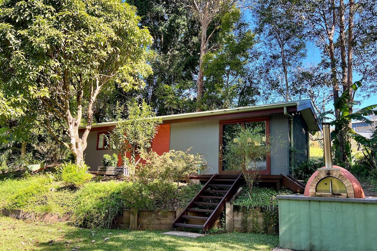 A charming standalone studio cottage is surrounded by lush greenery and trees. Steps lead up to the entrance of the cottage, which features large windows that invite natural light. A small outdoor area with a stone feature is also visible, enhancing the serene outdoor setting.