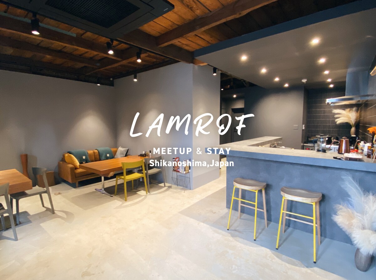 An open-plan living area features a modern kitchen island with bar stools and a dining table. Comfortable seating arrangements create a casual ambiance against grey walls and wooden beams. Soft lighting highlights the contemporary design, making it a welcoming space for social gatherings.