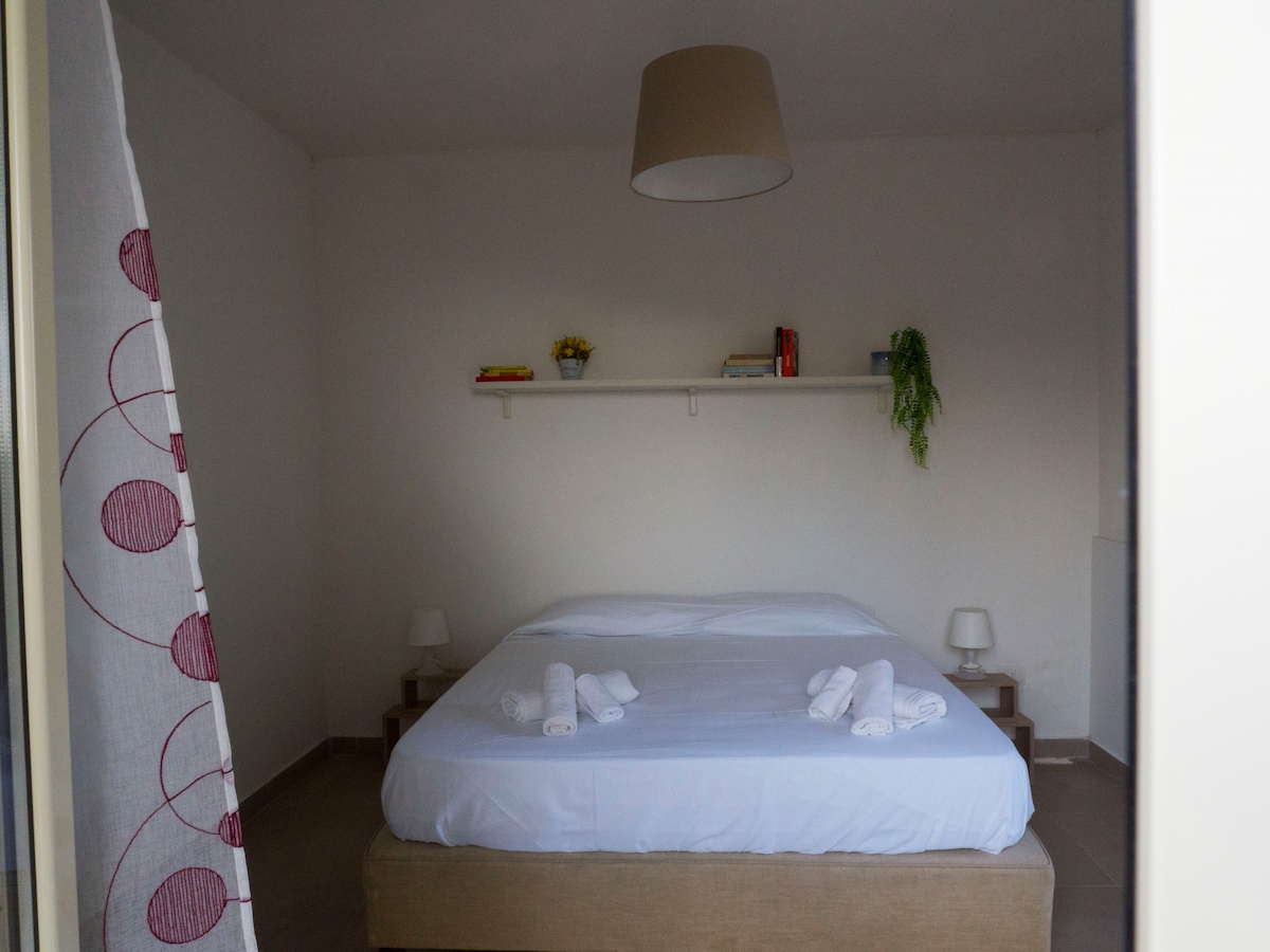 A spacious bedroom features a large bed with a crisp white duvet and neatly arranged towels. Two bedside tables with lamps flank the bed, while a shelf with decorative items and a hanging plant adorns the wall above. Soft, natural light enters through the window.