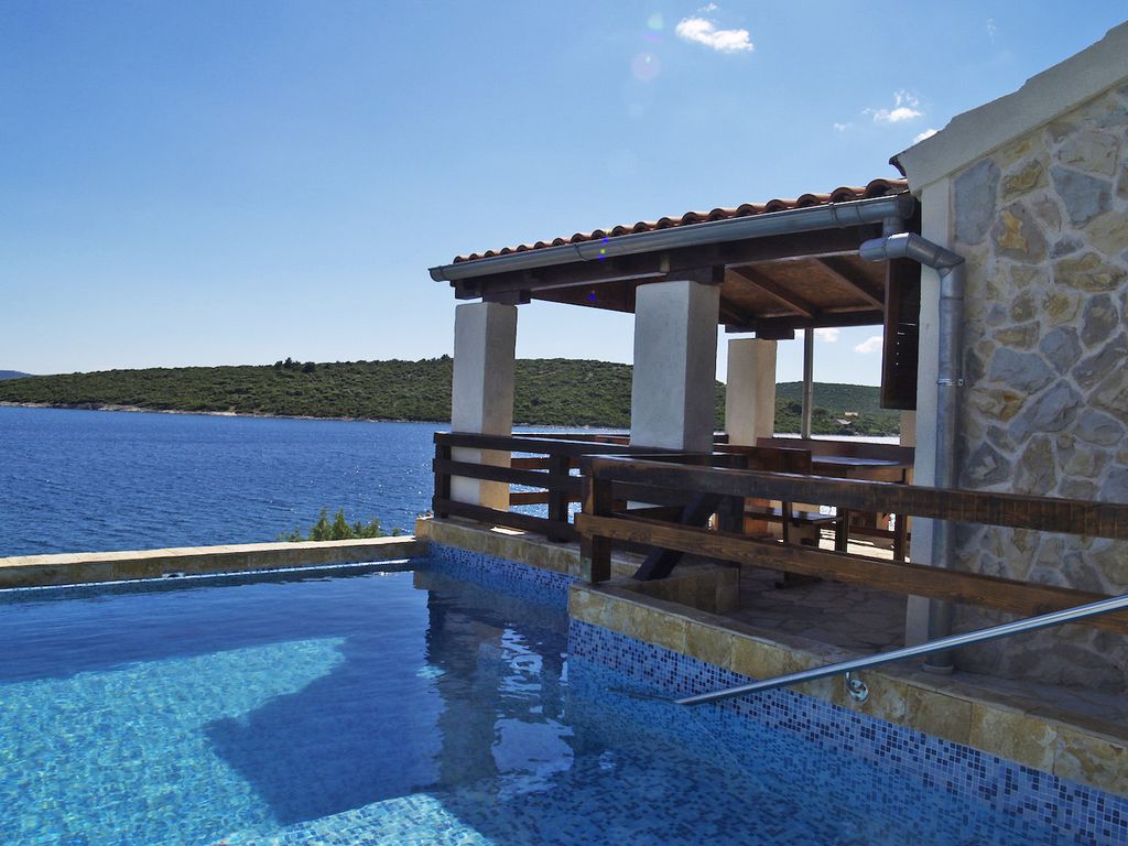 A private infinity pool overlooks the shimmering sea, complemented by a stone terrace with wooden railings. The rustic structure features an awning providing shade, with scenic views of the surrounding landscape visible across the water.