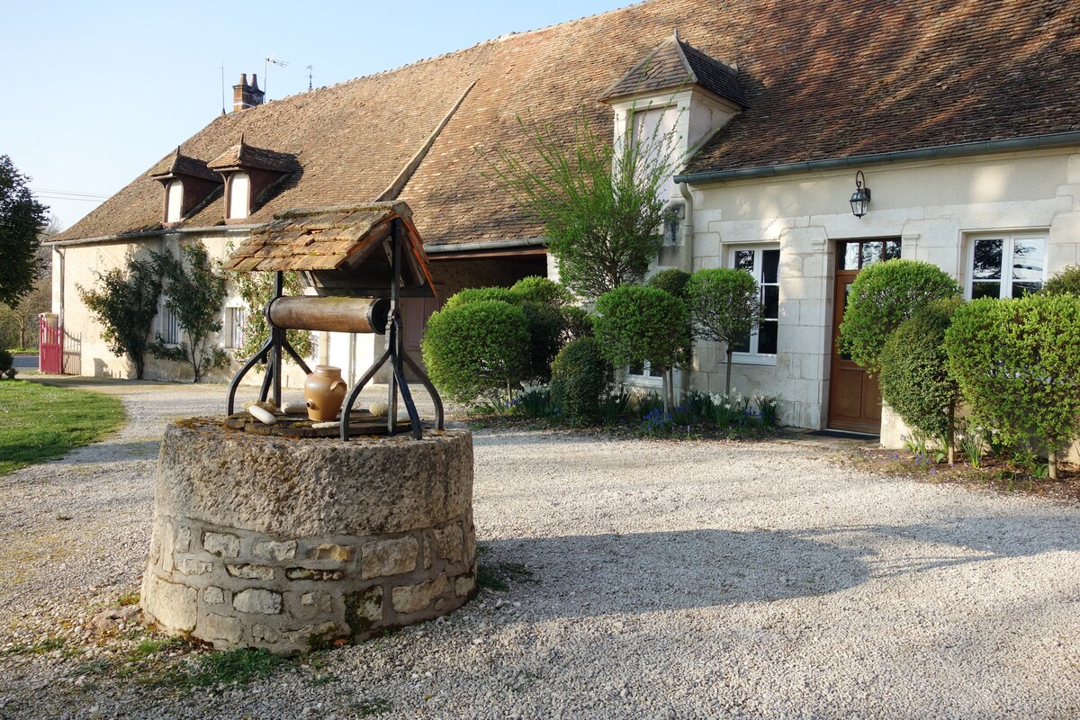 A historical building displays a rustic exterior with a traditional roof and multiple windows. A stone well is prominently featured in the foreground, surrounded by neatly trimmed bushes and a gravel pathway leading to the entrance.