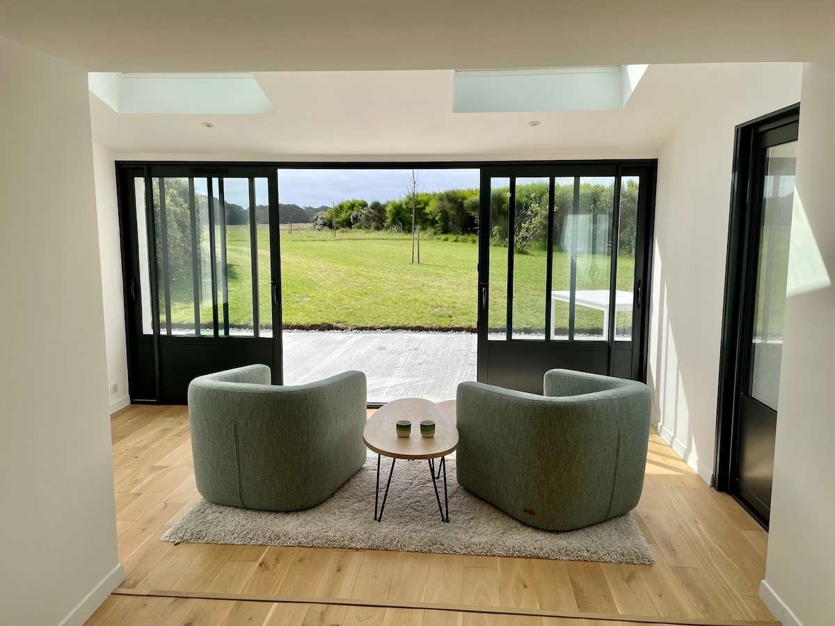 A bright seating area features two soft green chairs facing a small round table, positioned on a light wooden floor. Large glass doors slide open to reveal a view of a grassy landscape, creating a seamless connection between indoor and outdoor spaces.