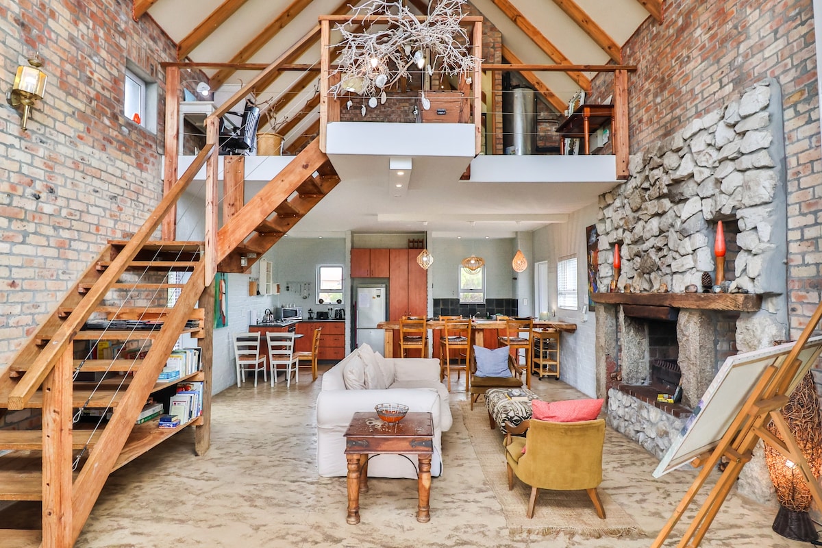 The open-plan living area features a high wooden ceiling and exposed brick walls, creating a spacious and airy feel. A large stone fireplace is located on one side, while a comfortable seating arrangement invites relaxation. A wooden staircase leads to a lofted area, enhancing the layout.