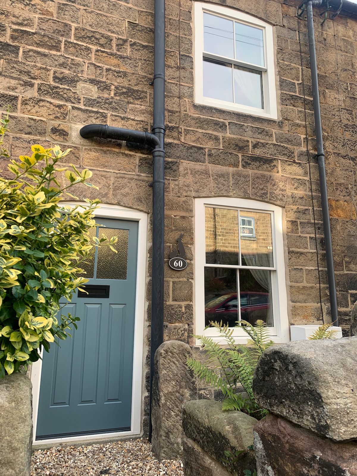 The entrance of a charming stone cottage features a blue panel door with a frosted glass window, framed by traditional stone walls. A gentle green foliage surrounds the entrance, enhancing the warm character of the home. Window boxes are adorned with rustic detailing.