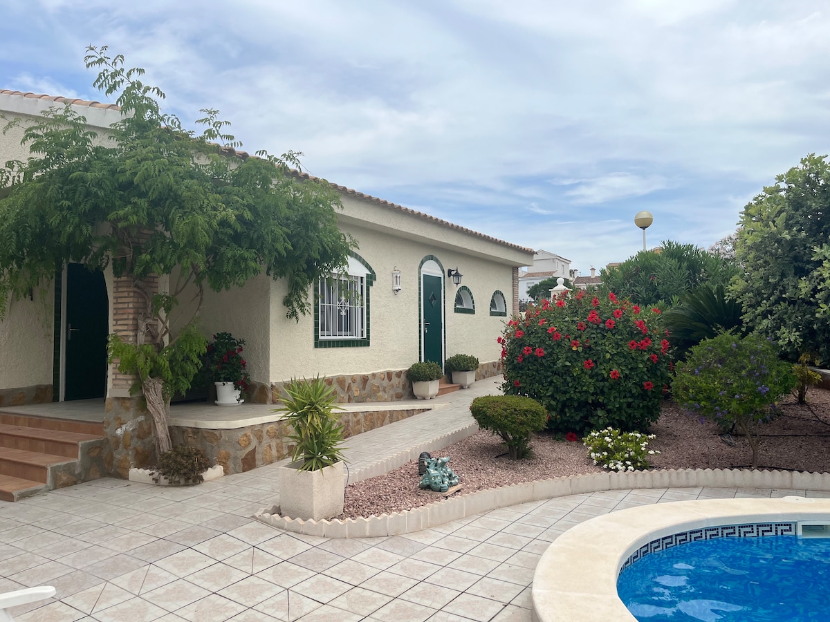 A charming exterior view of a three-bedroom accommodation is presented, surrounded by landscaped gardens featuring vibrant flowers and lush greenery. A swimming pool, framed by decorative tiles, complements the tranquil atmosphere, while a pathway leads to the inviting entrance with arched windows and doors.