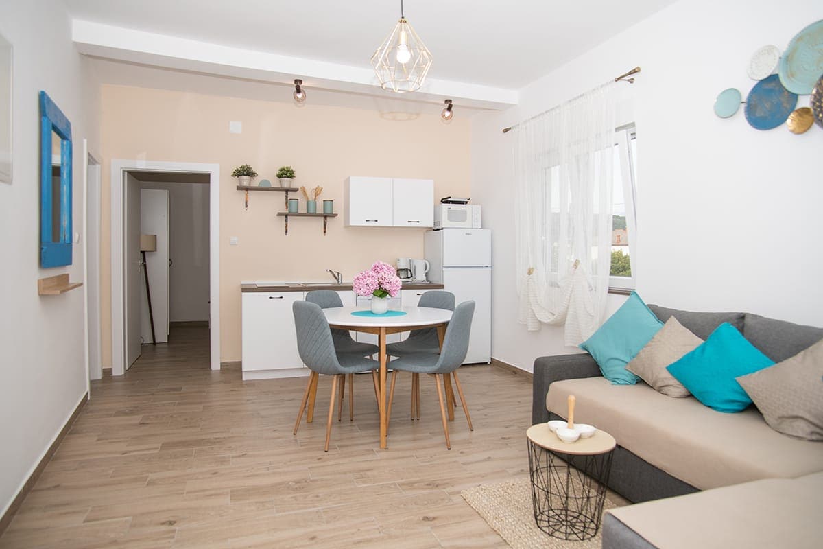 A cozy open-plan living area features a modern kitchen with white cabinetry and a dining table set for four. The sitting area includes a gray sofa with decorative pillows. Light wood flooring and soft white curtains enhance the space's welcoming ambiance.