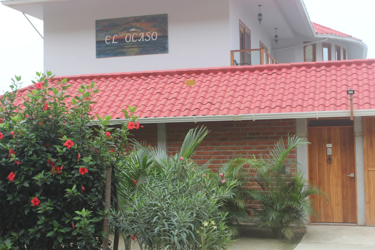 The exterior of the property showcases a red-tiled roof and a wooden door surrounded by vibrant greenery and tropical flowers. A sign reading 'El Ocaso' is displayed above the entrance, blending harmoniously with the natural surroundings.