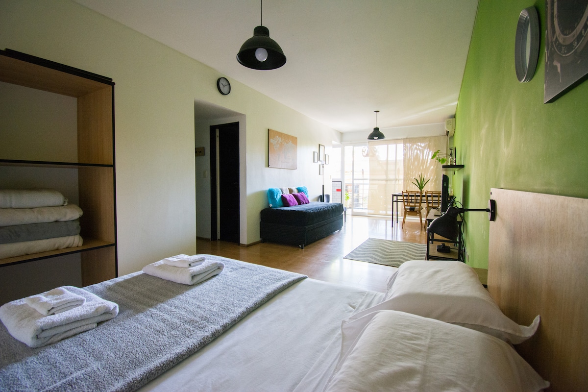 A studio apartment is displayed, featuring a spacious bed with neatly arranged towels. A cozy seating area with a dark sofa and colorful cushions is visible, along with a dining table. Light is let in through large glass doors, illuminating the green accent wall.