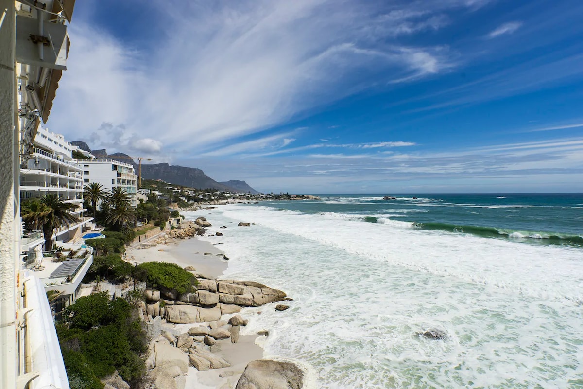 A panoramic view of the ocean showcases rolling waves meeting a sandy beach, bordered by rocky formations. Palm trees line the coast, and mountains are visible in the distance under a bright blue sky with scattered clouds.