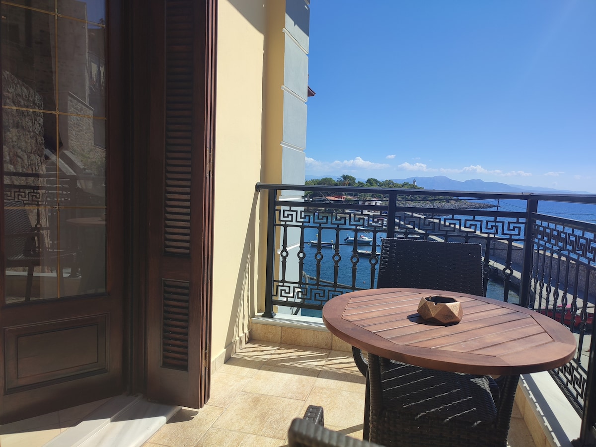 A charming balcony area offers a round wooden table surrounded by wicker chairs. The railing showcases a decorative design, and the scenic view includes the sparkling water of the Laconian Gulf and distant landforms under a clear blue sky.