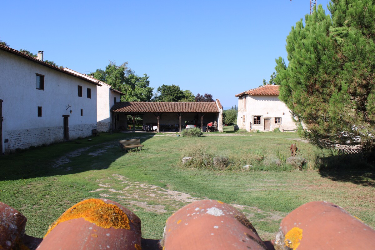 An expansive green lawn is framed by a rustic farmhouse and an adjacent building, both showcasing traditional architecture. A covered outdoor area is visible, providing shade, while trees dot the landscape in the background, contributing to a serene setting.