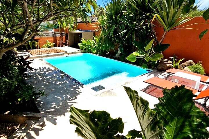 A private swimming pool is surrounded by lush tropical plants and trees, providing a sense of tranquility. Comfortable lounge chairs are positioned nearby for relaxation. The area features a mix of stone and concrete surfaces, with vibrant greenery enhancing the outdoor space.