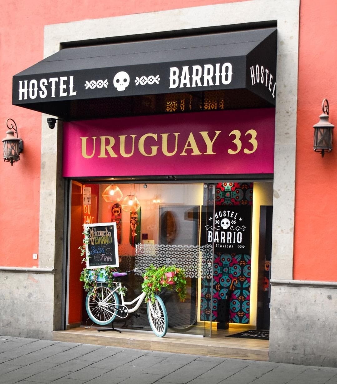 The entrance of the hostel displays a vibrant facade with a colorful sign featuring 'Hostel Barrio' and 'Uruguay 33.' A bicycle is adorned with plants, enhancing the inviting atmosphere. Wall decor in bright patterns provides a lively contrast to the building's exterior.