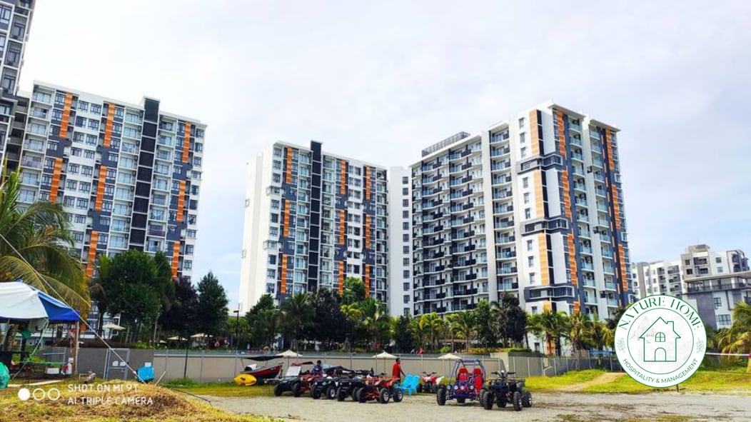 The image displays a modern multi-story building complex featuring a mix of colors on its exterior. In the foreground, various vehicles are parked on sandy ground, suggesting recreational activities. Lush greenery and palm trees can be seen surrounding the development, offering a tropical ambiance.