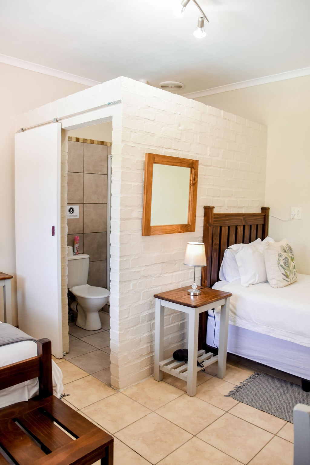 The interior space features two 3/4 beds separated by wooden nightstands. A mirror is mounted on a white brick wall, which partially encloses an en suite bathroom visible in the background. Natural light illuminates the room through a ceiling fixture.