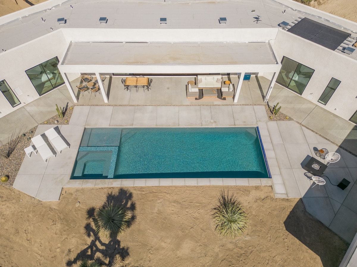 An aerial view showcases the modern home with a glistening pool surrounded by a spacious patio. Sun loungers are positioned nearby, and outdoor furniture is arranged under a shaded area, providing an inviting space for relaxation amidst the desert landscape.