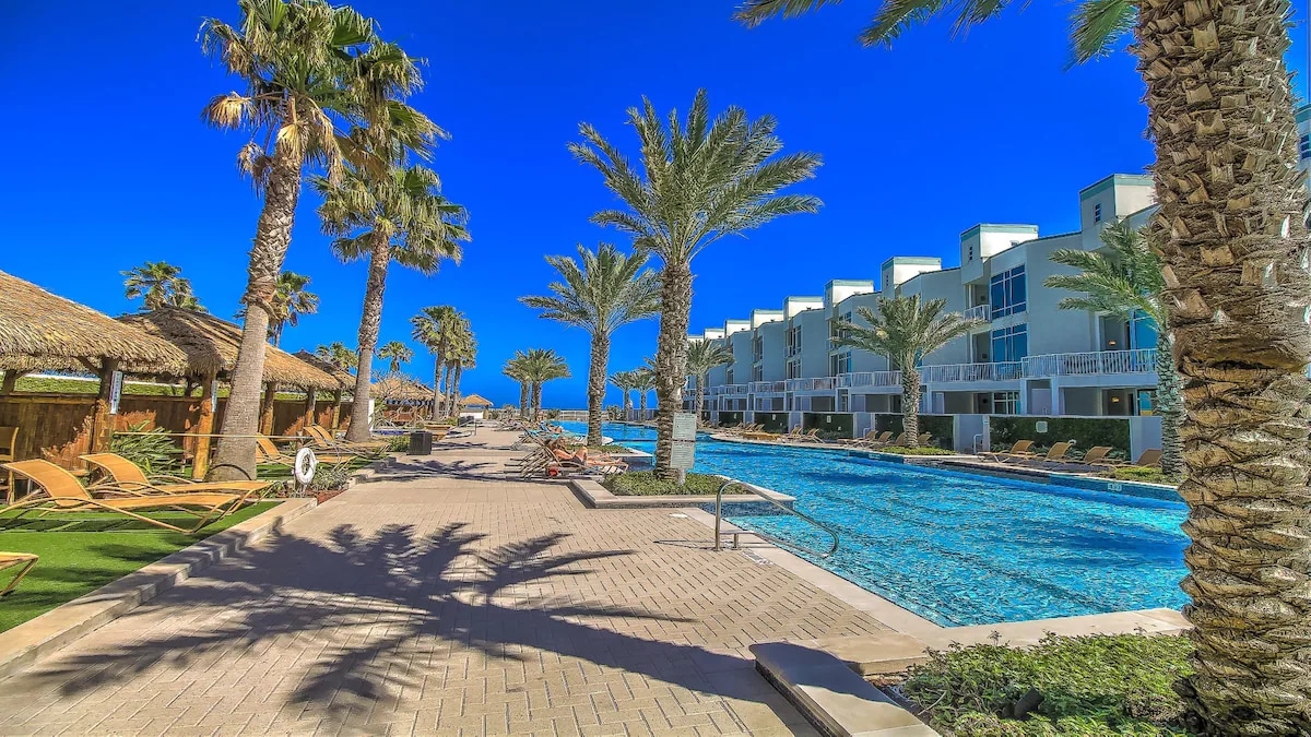 A landscaped outdoor area features a large pool surrounded by palm trees and lounge chairs. Thatched-roof cabanas provide additional shade, while a clear blue sky reflects the inviting atmosphere of the space. The modern building is visible in the background.