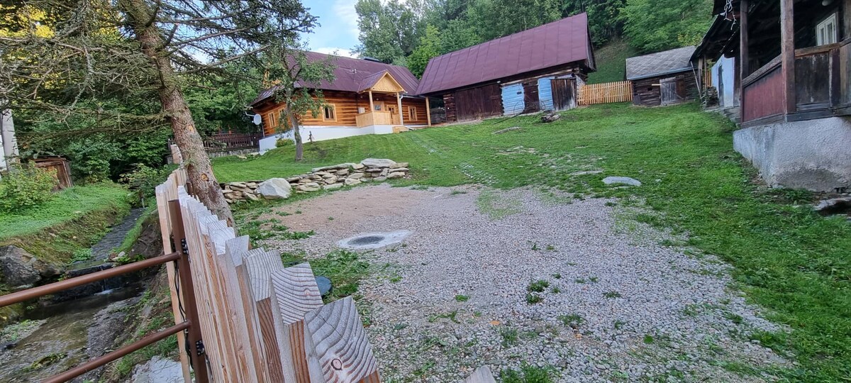 A spacious outdoor area features a gravel surface and a natural stone circle, surrounded by lush green grass. The property showcases multiple wooden structures, including a main cabin with a sloped roof and an adjacent smaller building. A tree line and hillside provide a serene backdrop.