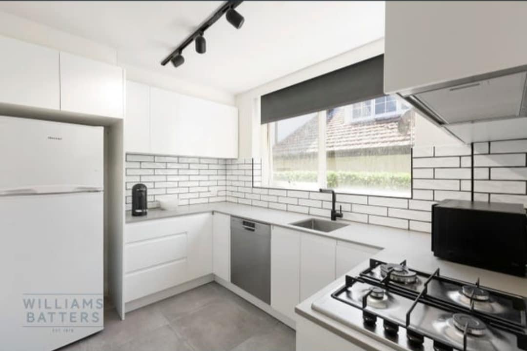 A modern kitchen is displayed, featuring white cabinetry and a gray tile floor. Natural light flows through a large window, illuminating the stainless steel appliances, including a dishwasher and a gas stove. Black window shades provide a sleek contrast against the bright decor.