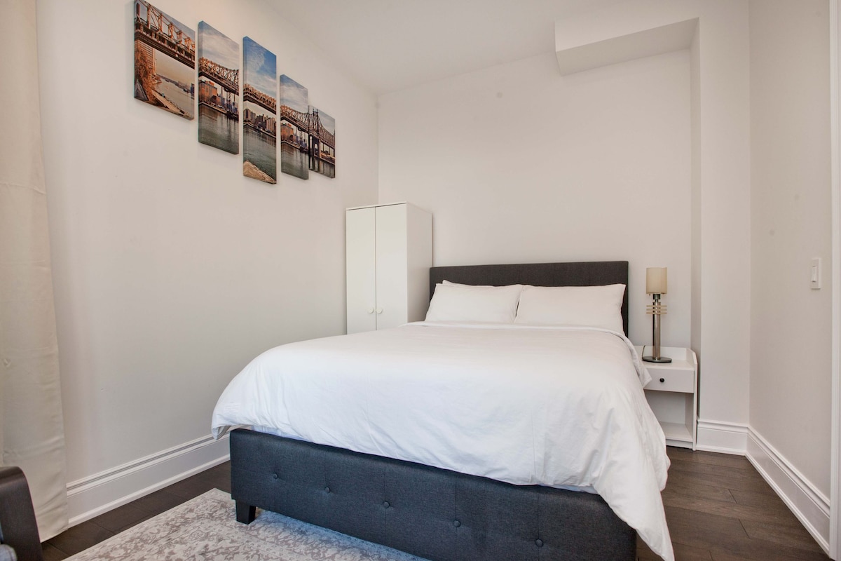A bright sleeping area features a spacious bed with white bedding, accompanied by a bedside table with a lamp. The room is adorned with a set of five landscape photographs on the wall, and a wardrobe stands nearby, providing ample storage.
