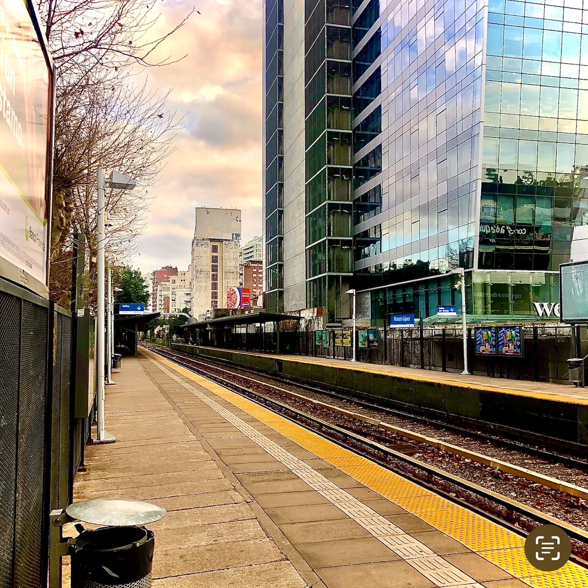 A clean and spacious train platform is visible, lined with yellow tactile tiles. Modern buildings reflect in the glass of a nearby structure, with hints of urban life in the background. The tracks are empty, and the surrounding area is well-maintained.