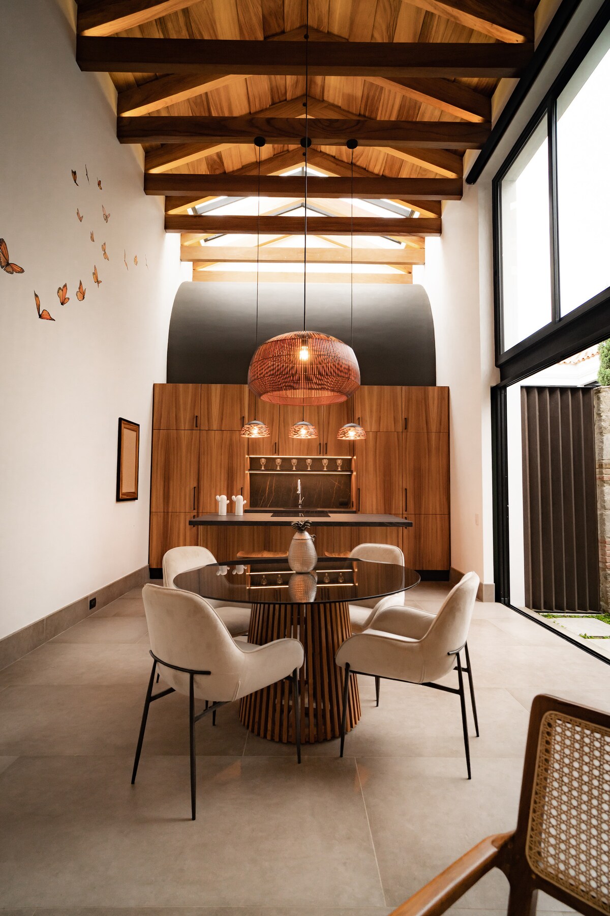 A dining area is highlighted by a round table surrounded by four armchairs, set under a high wood-beamed ceiling. A large pendant light hangs above the table, and a wooden cabinetry provides a warm contrast to the light-colored walls and floor.