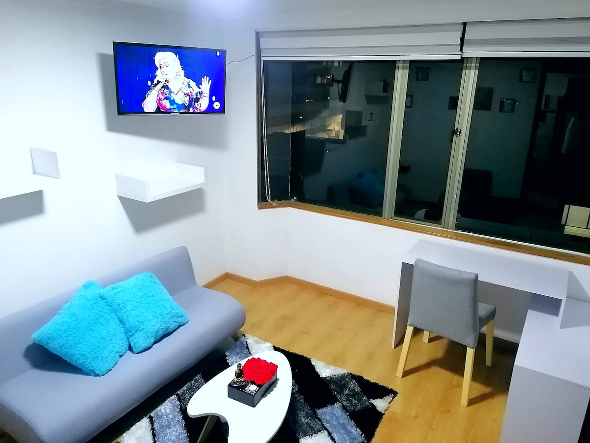 A well-lit living area is showcased with a gray sofa adorned with bright blue pillows. A sleek white coffee table sits in front of the sofa, and a television is mounted on the wall, displaying a performance. Large windows allow for a view of the city.