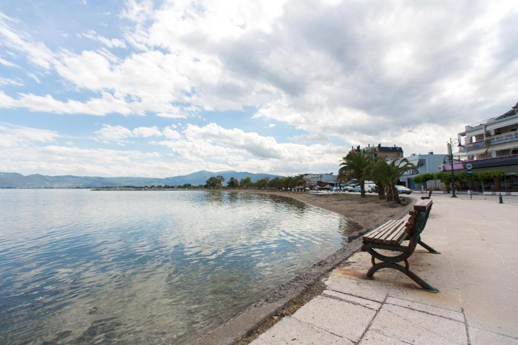 A peaceful waterfront view is characterized by calm waters reflecting the sky. A wooden bench is positioned along a paved walkway, bordered by palm trees. The horizon features distant mountains, while nearby buildings provide a glimpse of local architecture.