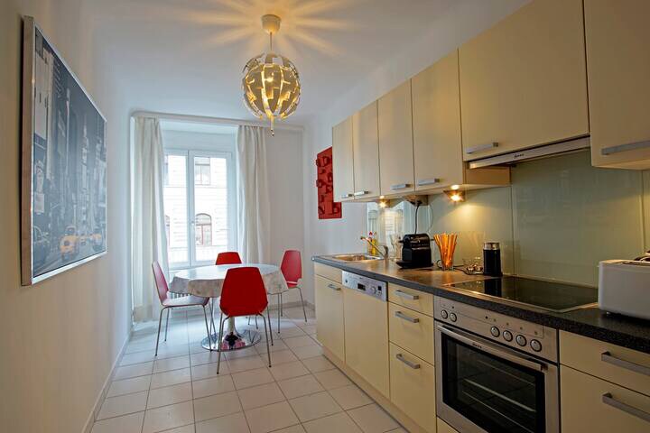 A light-filled kitchen features modern cabinetry and stainless steel appliances. A round dining table with red chairs is positioned near a window, complemented by a pendant light overhead. A piece of art is mounted on the wall, adding a contemporary touch.