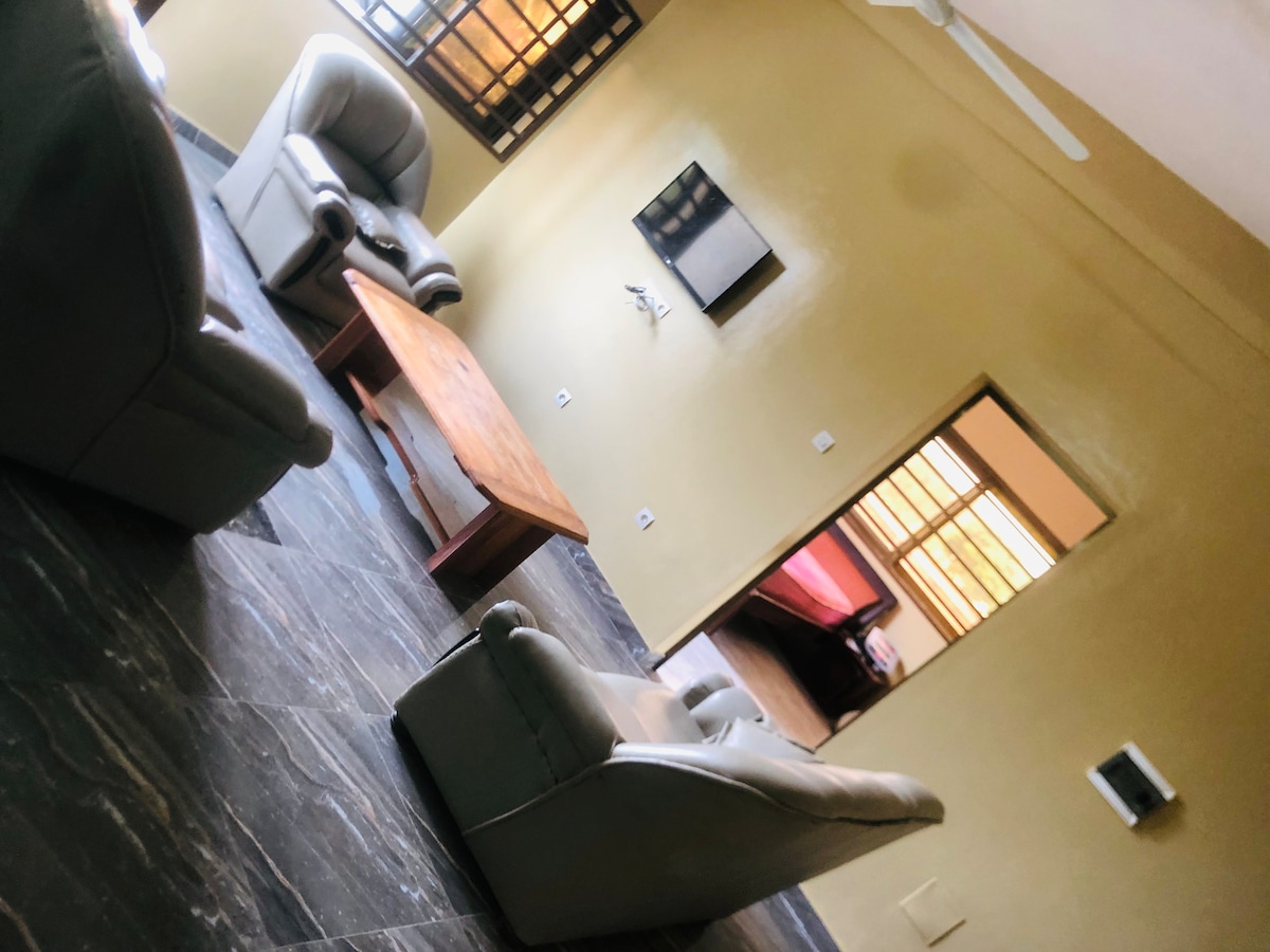 A spacious living area displays two comfortable armchairs and a wooden coffee table set on dark tiled flooring. A wall-mounted TV is visible, and a doorway leads to a softly lit bedroom beyond. Large windows allow natural light to fill the room.