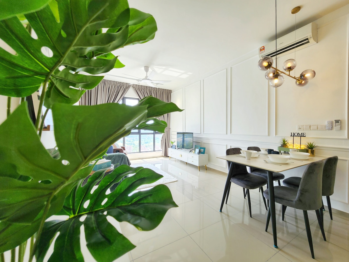 A dining area is visible with a modern table set for four, complemented by plush chairs. A large window allows natural light to fill the space, while a stylish plant adds a touch of greenery. A television is positioned against a light-colored wall.