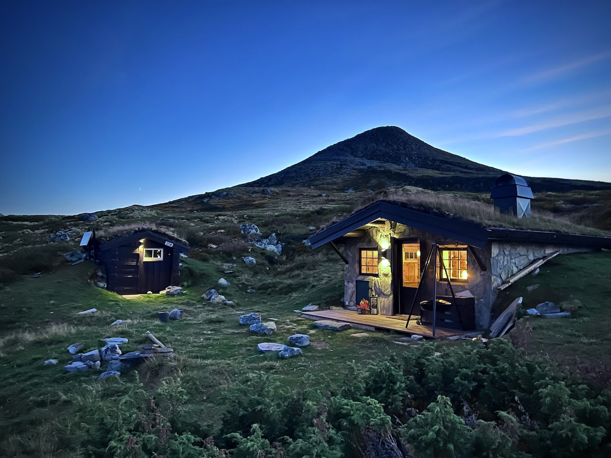 A scenic mountain landscape at dusk shows a rustic cabin with warm light glowing from its windows. An adjacent outbuilding is softly illuminated, set against the backdrop of a prominent peak. Natural greenery and rocky features surround the structures, enhancing the serene atmosphere.