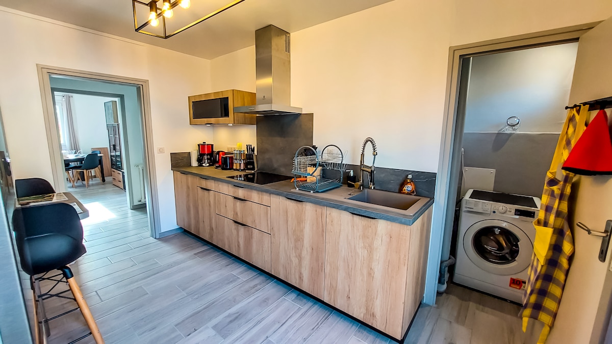 A modern kitchen is featured, showcasing wooden cabinetry and a sleek gray countertop. Stainless steel appliances, including a range hood, are visible. A washing machine occupies a corner, and bar stools are positioned at the counter, providing a functional cooking area.