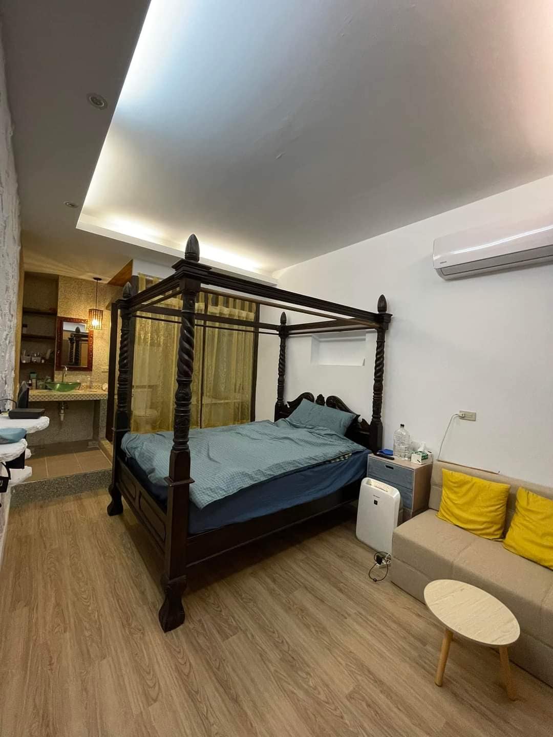 A spacious room features a dark wooden four-poster bed draped with light fabric. A small sofa and a round side table are arranged nearby. The space is illuminated by soft lighting, creating a calming atmosphere. A kitchenette area is visible in the background.