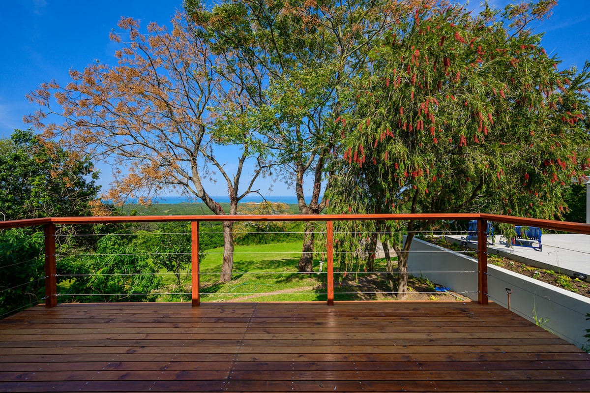 A wooden deck provides a spacious outdoor area framed by lush greenery and flowering trees. The expansive view features rolling lawns and a glimpse of the sea in the distance under a clear blue sky.