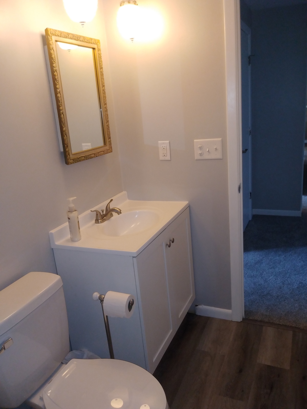 A clean and simple bathroom features a white vanity with a gold-finished faucet and a round mirror framed in a gold design. A white toilet sits beside the vanity, and a roll of toilet paper is positioned within reach. The flooring is a light wood material.