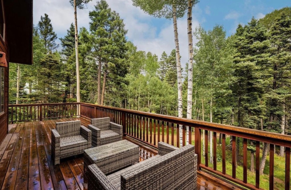 A spacious wooden deck features a set of wicker patio chairs arranged for relaxation. Lush trees surround the area, providing a natural privacy screen. The deck is positioned to offer a tranquil view of the forest, inviting guests to unwind in the fresh mountain air.